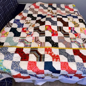 May include: A white quilt with a patchwork design featuring various colors and patterns, including red, blue, green, and brown. The quilt is made up of squares and rectangles, and the patterns are arranged in a geometric design. The quilt is laid out on a bed, and a measuring tape is visible at the bottom of the image.