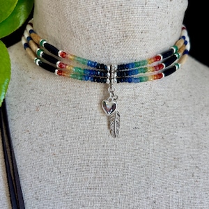 May include: A three-strand choker necklace featuring alternating segments of tan wood, black, and multi-colored gemstone beads in a rainbow gradient. The necklace includes a silver-toned metal connector with a dangling heart and feather charm.