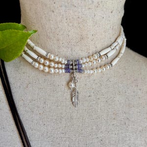 May include: A multi-strand choker necklace featuring white cylindrical beads, cream-colored pearls, and small purple accent beads. The center includes a silver-toned metal connector with a dangling silver feather charm and a single white pearl.