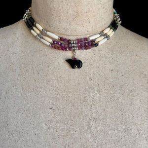 May include: A multi-strand necklace featuring white, black, and purple beads. The necklace has a black bear pendant with silver accents. The necklace is displayed on a mannequin.