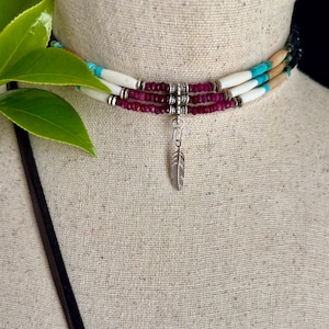 May include: A multi-strand choker necklace with turquoise, white, and maroon beads. The necklace has a silver feather pendant. The necklace is displayed on a mannequin.