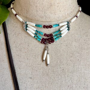 May include: A multi-strand choker necklace featuring white cylindrical beads, turquoise-coloured spacer beads, and dark red faceted beads. The design includes silver-toned metal accents and two dangling white cylindrical bead charms at the centre.