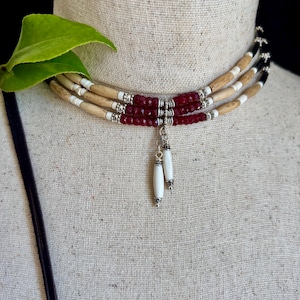 May include: A three-strand choker necklace featuring light tan cylindrical beads, small white spacers, and deep red faceted beads. The centre includes silver-toned metal accents and two dangling white cylindrical pendants with silver-toned caps.