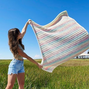 May include: A person holding a large, striped crochet blanket in a field of tall grass. The blanket is made with a variety of pastel colors, including pink, blue, and green.