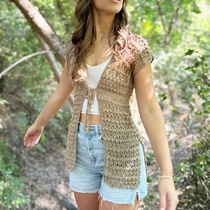 May include: A woman wearing a light brown crochet cardigan with a tie front and short sleeves. She is wearing blue denim shorts and has long brown hair.