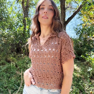 May include: A short-sleeved, brown crochet top with a collared neckline and open weave design. The top is paired with light blue denim shorts. The model is standing outdoors with a natural green background.