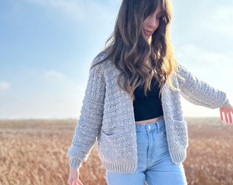 May include: A woman wearing a light gray crocheted cardigan with pockets and blue jeans stands on a wooden platform in a field of tall grass.