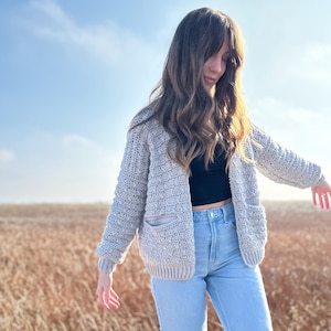 Peut inclure: Une femme portant un cardigan en crochet gris clair avec des poches et un jean bleu se tient sur une plateforme en bois dans un champ d'herbe haute.