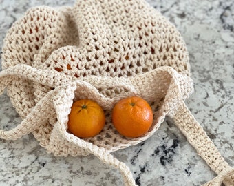 May include: A white crocheted tote bag with two orange mandarins inside. The bag is lying on a light gray countertop.
