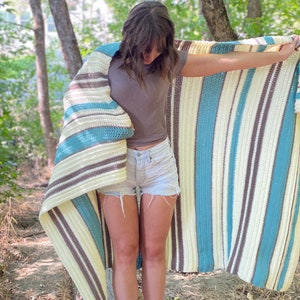 May include: A striped crocheted throw blanket in shades of brown, cream, and teal. The blanket is draped over a person's shoulders.