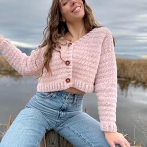 May include: A light pink, cropped cardigan with three brown buttons. The cardigan is paired with light blue jeans. The outfit is worn outdoors, with a background of water and reeds.