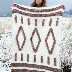 May include: A white crocheted throw blanket with a brown fringe and a diamond pattern. The blanket is being held by a person in a snowy field.