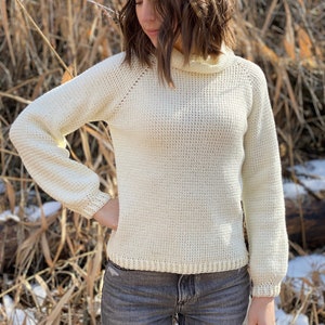 May include: A white crocheted sweater with a mock neck and long sleeves. The sweater has a textured stitch pattern and a ribbed hem.