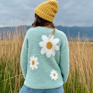 Pode incluir: Um suéter azul claro com grandes aplicações de margaridas brancas e centros amarelos. O suéter é combinado com um gorro amarelo mostarda e jeans azuis. A pessoa está em um campo de grama alta, com montanhas ao fundo.
