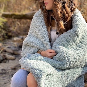 Puede incluir: Una mujer sentada al aire libre envuelta en una suave manta peluda de color azul claro. Lleva pantalones vaqueros azules y una camisa a cuadros.