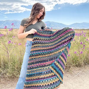 Peut inclure: Une couverture au crochet faite à la main, aux couleurs vives, avec des rayures bleues, roses, vertes et jaunes. La couverture est présentée sur fond de champ de fleurs mauves, de montagnes et de ciel nuageux.