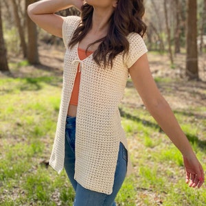 May include: A white crochet cardigan with a tie at the neckline. The cardigan has a loose fit and is open in the front. The model is wearing the cardigan over a coral tank top and blue jeans.