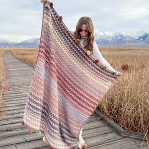 以下が含まれることがあります： 縁にポンポンが付いた、シェブロン柄のマルチカラーのクロシェ編み毛布。毛布は木製の歩道にいる人によって持たれています。