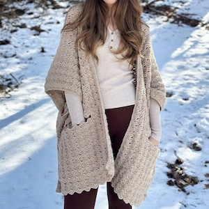 May include: A beige crochet shawl with pockets, worn over a white long-sleeved shirt and brown pants. The person is standing in a snowy outdoor setting.