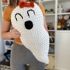 May include: A white crocheted ghost with black eyes and a black mouth. The ghost has an orange bow on top of its head.