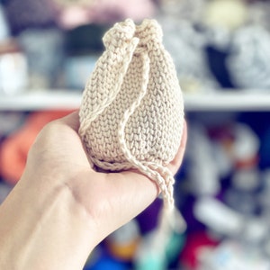 May include: A small, off-white knitted drawstring bag with a gathered top and a long, knotted cord.