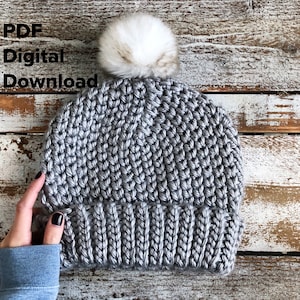 Puede incluir: Gorro de punto de ganchillo gris con un pompón de piel sintética blanca. El gorro tiene un borde de canalé y un patrón de punto texturizado. Descarga digital PDF.