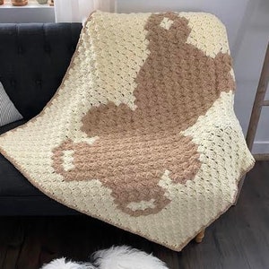 Pode incluir: Uma manta de croch&ecirc; branca e marrom com um padr&atilde;o geom&eacute;trico. A manta est&aacute; drapeada sobre um sof&aacute; preto e um c&atilde;o branco est&aacute; deitado no ch&atilde;o em frente ao sof&aacute;.