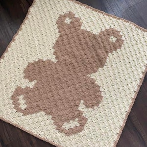 Pode incluir: Uma manta de croch&ecirc; branca com uma silhueta de urso de croch&ecirc; marrom no centro.