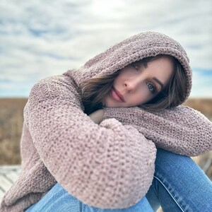 May include: A woman wearing a soft, pink hooded sweater and blue jeans, sitting with her arms crossed and looking at the camera.