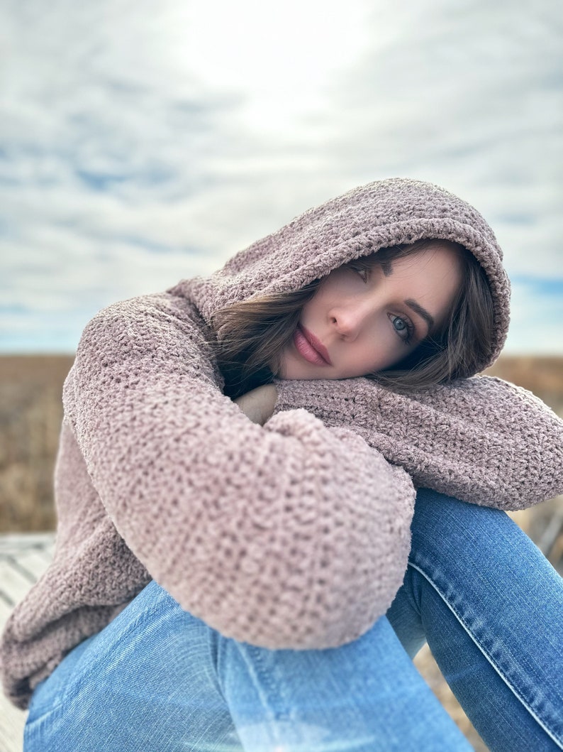 May include: A woman wearing a soft, pink hooded sweater and blue jeans, sitting with her arms crossed and looking at the camera.