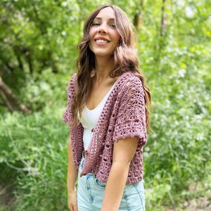 May include: A mauve crochet shrug with short sleeves. The open-weave design is worn over a white top and light blue denim shorts. The background is a lush green forest.
