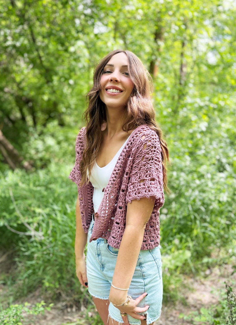 May include: A mauve crochet shrug with short sleeves. The open-weave design is worn over a white top and light blue denim shorts. The background is a lush green forest.