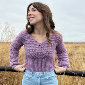 Peut inclure: Un pull lilas à manches longues avec un col en V et un motif en tricot texturé. Le pull a un ourlet et des poignets côtelés. La personne porte un jean bleu clair. L'arrière-plan présente un champ d'herbe sèche et un ciel nuageux.
