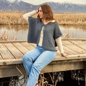 Puede incluir: Un poncho de ganchillo azul grisáceo con cierre de lazo, sobre una camiseta crema y vaqueros azules. El poncho tiene un patrón texturizado. El fondo muestra un muelle de madera, agua y montañas.