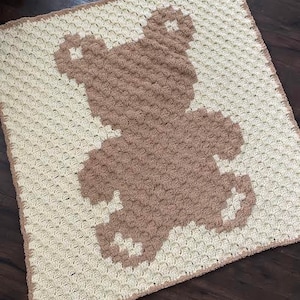 Pode incluir: Uma manta de croch&ecirc; com um desenho de urso castanho. A manta &eacute; feita com fio branco e castanho e tem um padr&atilde;o texturizado de croch&ecirc;.
