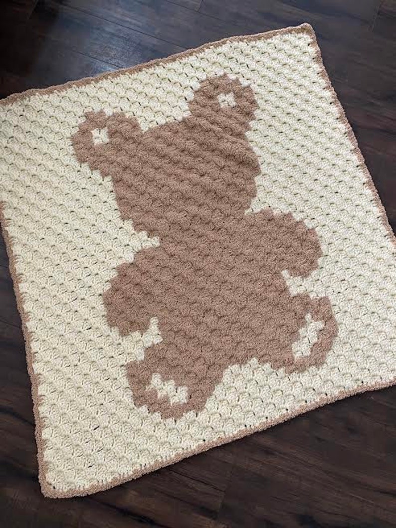 Pode incluir: Uma manta de croch&ecirc; com um desenho de urso castanho. A manta &eacute; feita com fio branco e castanho e tem um padr&atilde;o texturizado de croch&ecirc;.
