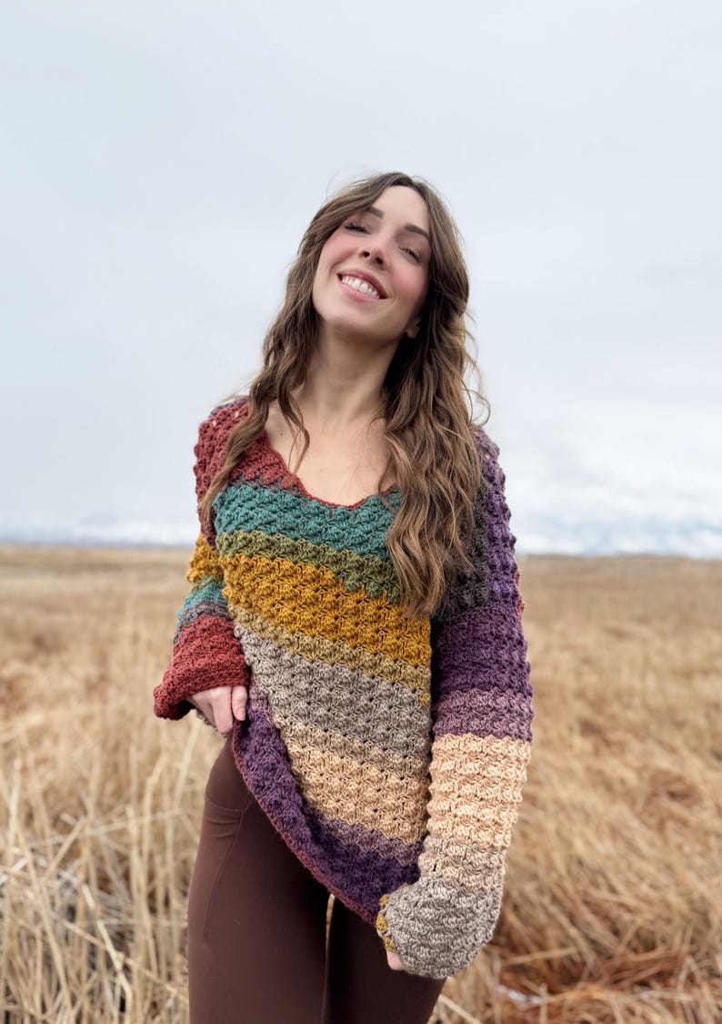 May include: A multi-coloured crochet sweater with a v-neck and long sleeves. The sweater is made in a variety of shades of brown, green, yellow, and purple.