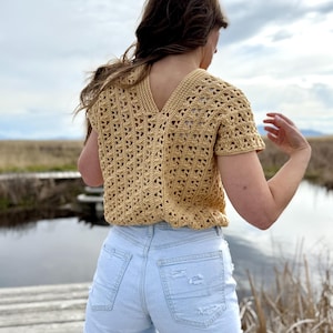 May include: A short-sleeved, light yellow crochet top with a V-neck. The top is paired with light blue denim shorts. The image is taken outdoors with a body of water and cloudy sky in the background.