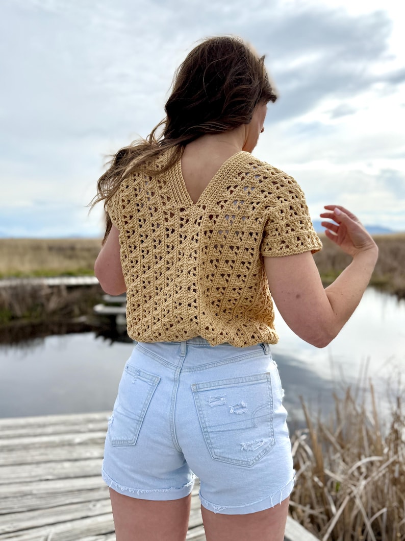 May include: A short-sleeved, light yellow crochet top with a V-neck. The top is paired with light blue denim shorts. The image is taken outdoors with a body of water and cloudy sky in the background.