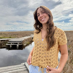 May include: A mustard yellow crochet top with a V-neck and short sleeves. The top has a drawstring at the waist and a geometric openwork pattern. The person is wearing light blue jeans and is standing outside near a body of water.