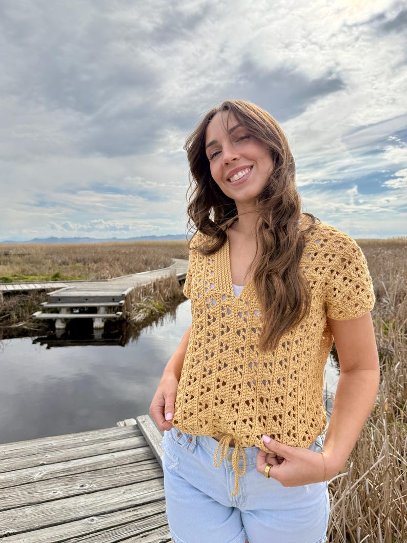 May include: A mustard yellow crochet top with a V-neck and short sleeves. The top has a drawstring at the waist and a geometric openwork pattern. The person is wearing light blue jeans and is standing outside near a body of water.