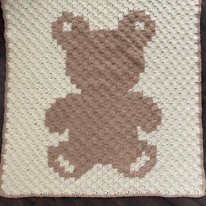 Pode incluir: Um cobertor de beb&eacute; de croch&ecirc; com uma silhueta de urso castanho no centro. O cobertor &eacute; feito com fio branco e o urso &eacute; feito com fio castanho.