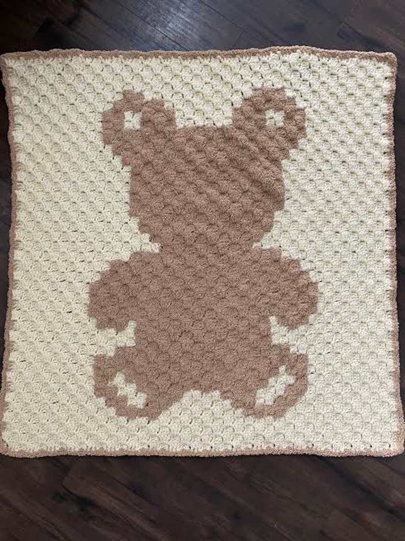 Pode incluir: Um cobertor de beb&eacute; de croch&ecirc; com uma silhueta de urso castanho no centro. O cobertor &eacute; feito com fio branco e o urso &eacute; feito com fio castanho.