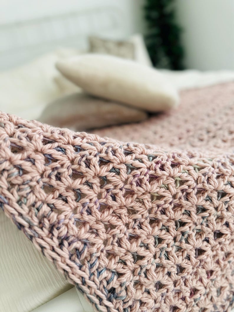May include: A close-up of a crocheted throw blanket in a soft pink and purple color scheme. The blanket is made with a delicate stitch pattern and has a soft, textured feel.