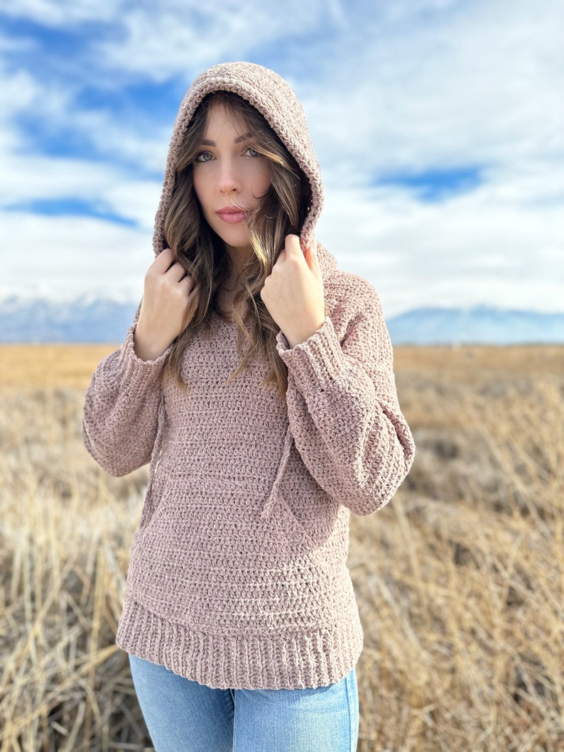 May include: A mauve crocheted hooded sweater with a drawstring. The sweater is worn by a person standing in a field of dry grass.