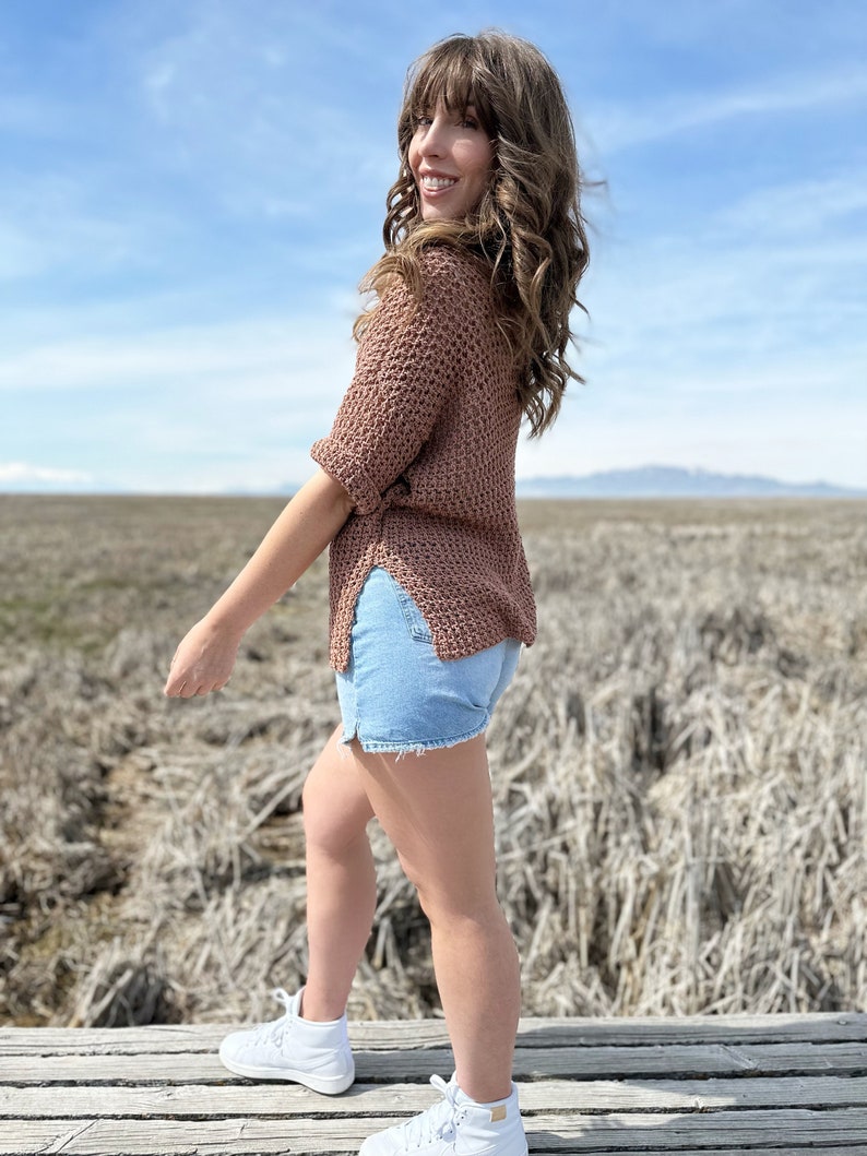Puede incluir: Una mujer con un su&eacute;ter de punto marr&oacute;n, pantalones cortos de mezclilla y zapatillas blancas. Est&aacute; de pie en una plataforma de madera con un campo de hierba seca en el fondo.