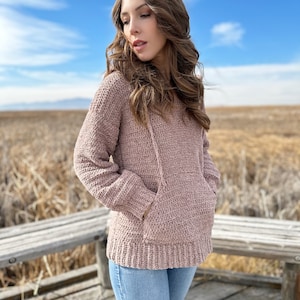 May include: A woman wearing a mauve crocheted hooded sweater with a pocket. She is standing on a wooden platform with a field of dry grass in the background.