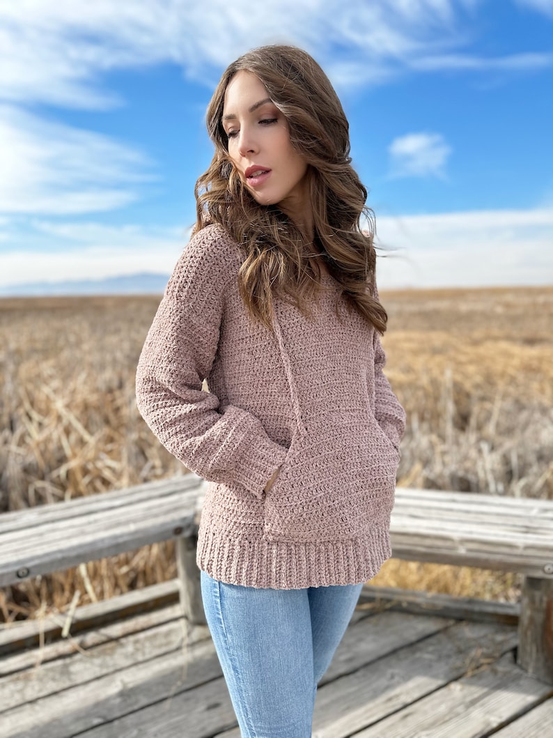 May include: A woman wearing a mauve crocheted hooded sweater with a pocket. She is standing on a wooden platform with a field of dry grass in the background.