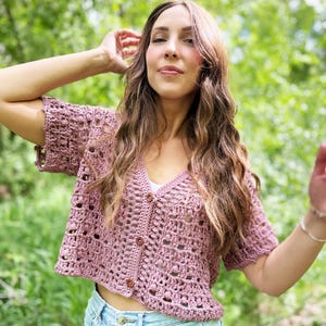 Könnte beinhalten: Ein mauvefarbener Häkelcardigan mit kurzen Ärmeln, V-Ausschnitt und Holzknöpfen. Das offene Webmuster ist sichtbar. Der Cardigan wird mit hellblauen Jeans getragen.