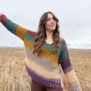 May include: A woman wearing a multi-coloured crochet sweater with a diagonal stripe pattern. The sweater is made with a textured stitch and features shades of brown, green, purple, and yellow.